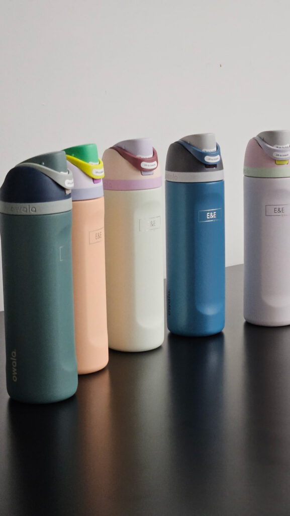 Five engraved water bottles in green, peach, cream, blue and lavender standing in a row on a dark tabletop