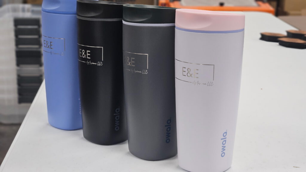 Close-up of four tumblers in blue, black, gray and white, each engraved with a boxed E&E logo and Ag Insurance LLC in script