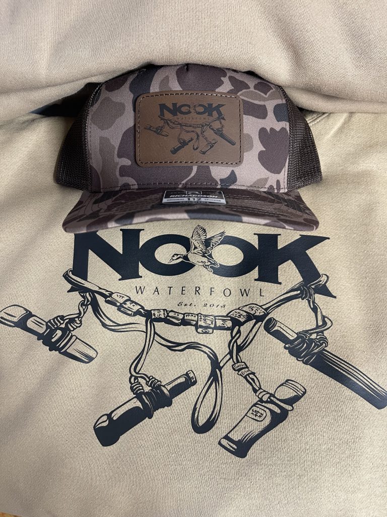Dark camo trucker hat with an engraved Nook Waterfowl leatherette patch, resting on a cream tee printed with the Nook Waterfowl logo