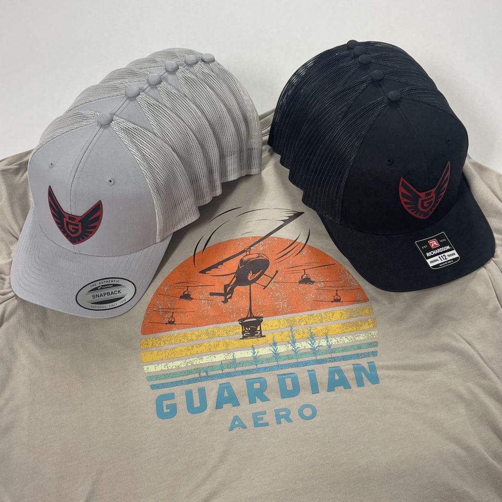Gray and black trucker hats with red and black Guardian Aero winged-G patches, on a sand tee printed with a retro sunset design of a helicopter over rows of crops and Guardian Aero in blue