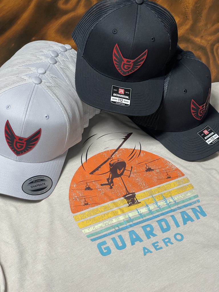Gray and black Guardian Aero patch hats piled on the sand tee with the sunset helicopter design, against a wood-grain background
