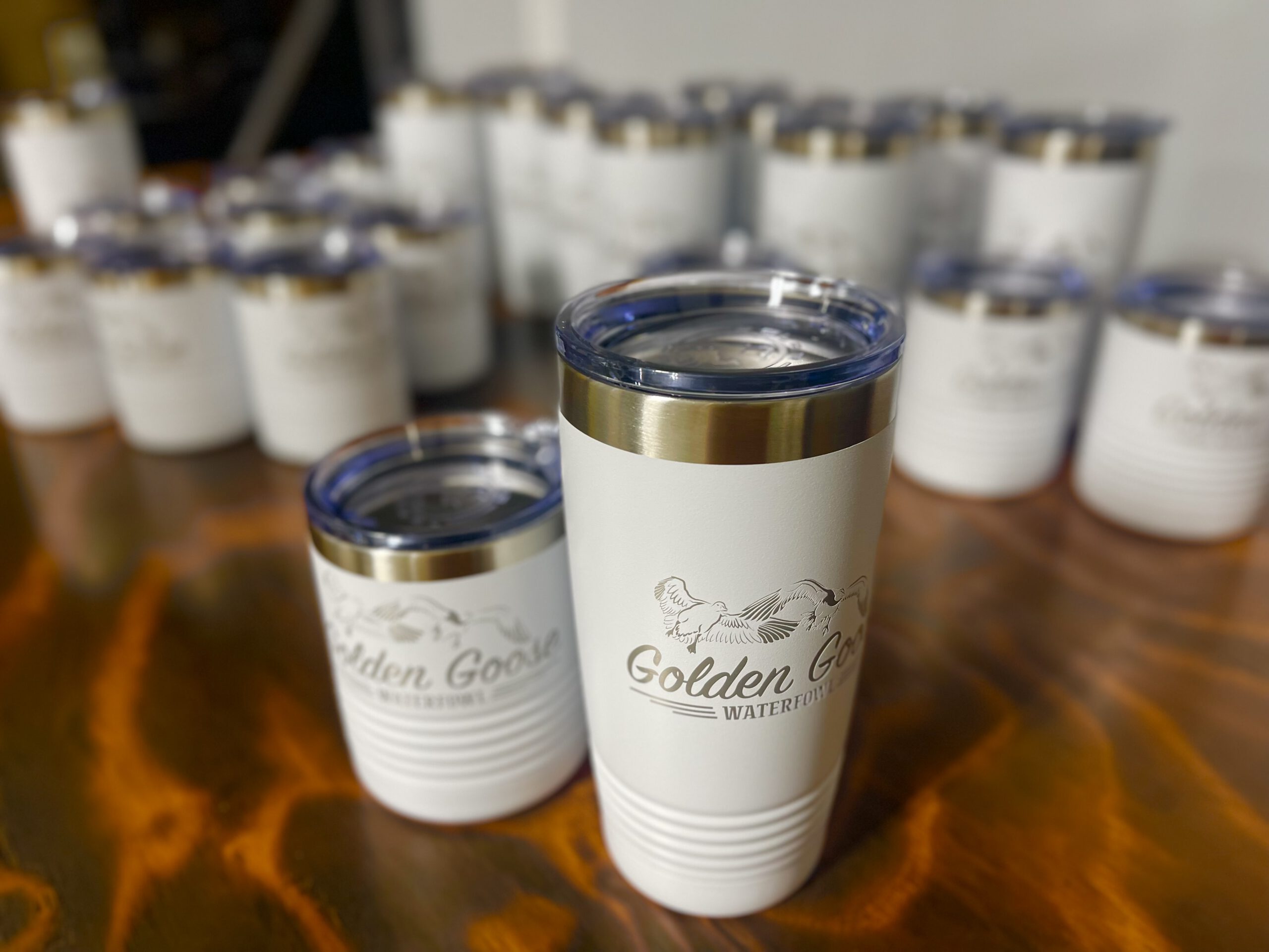White tumblers engraved with the Golden Goose Waterfowl logo, two in front and more lined up behind