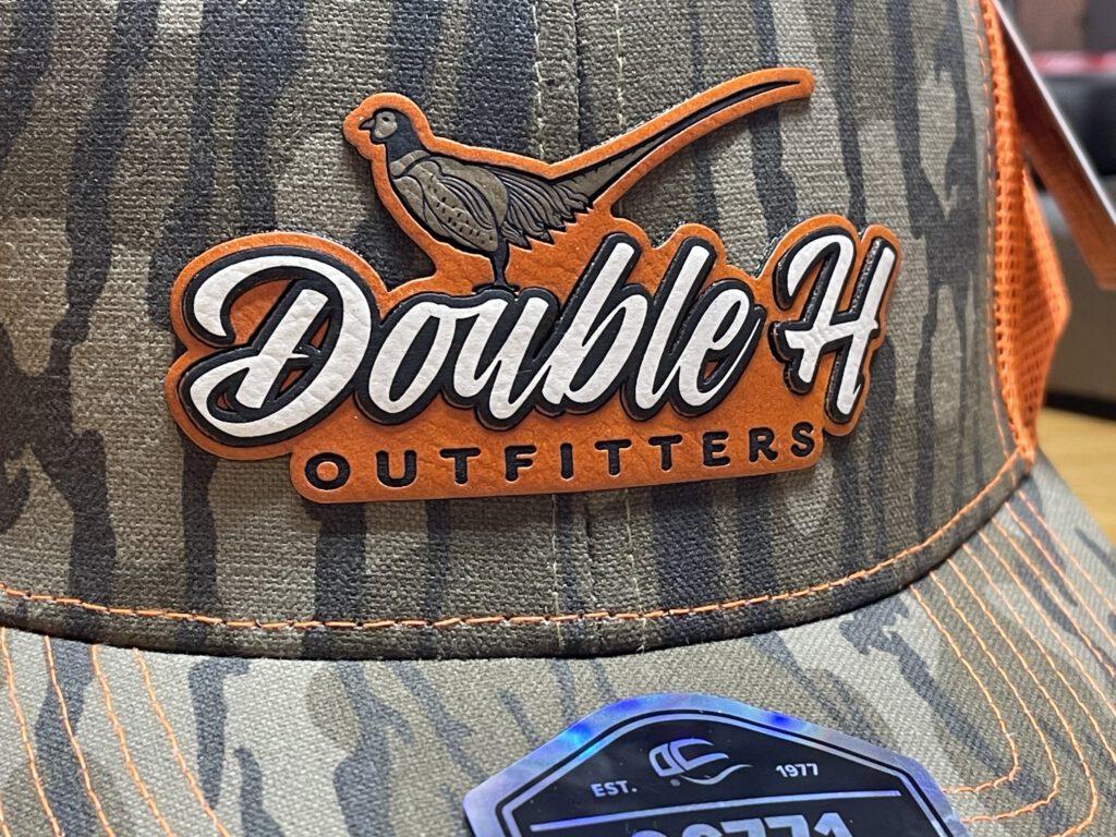 Close-up of a Double H Outfitters inlaid leatherette patch on a camo hat: a pheasant above the script logo on an orange backing
