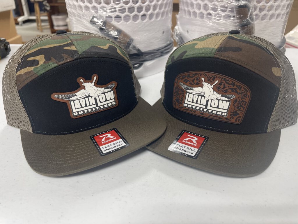 Two camo and black snapback hats with the same two Layin' Low Outfitters patch styles