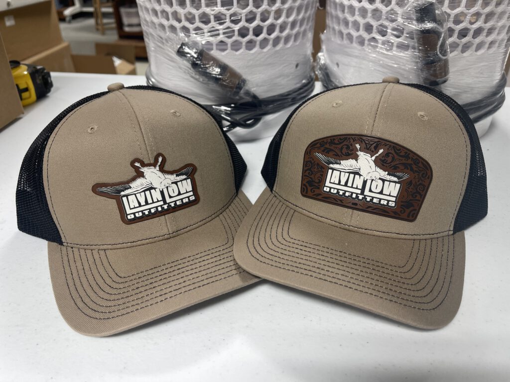 Two khaki and black trucker hats with Layin' Low Outfitters patches: one cut to the shape of the logo, one a layered patch with a tooled-leather pattern