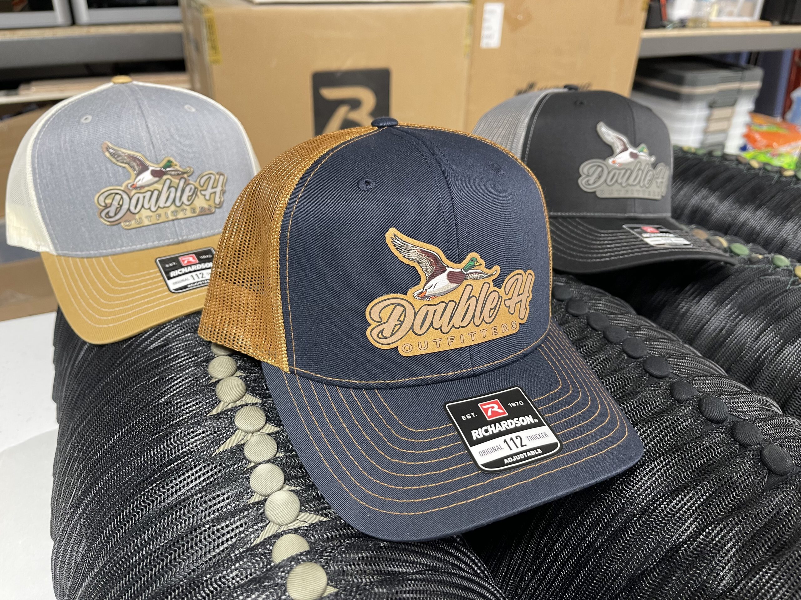 Three trucker hats with full-color Double H Outfitters patches showing the mallard script logo, the front one navy and brown