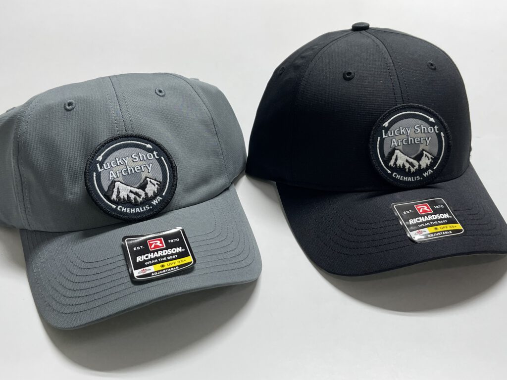 Gray and black hats with round Lucky Shot Archery patches showing mountains and the words Chehalis, WA