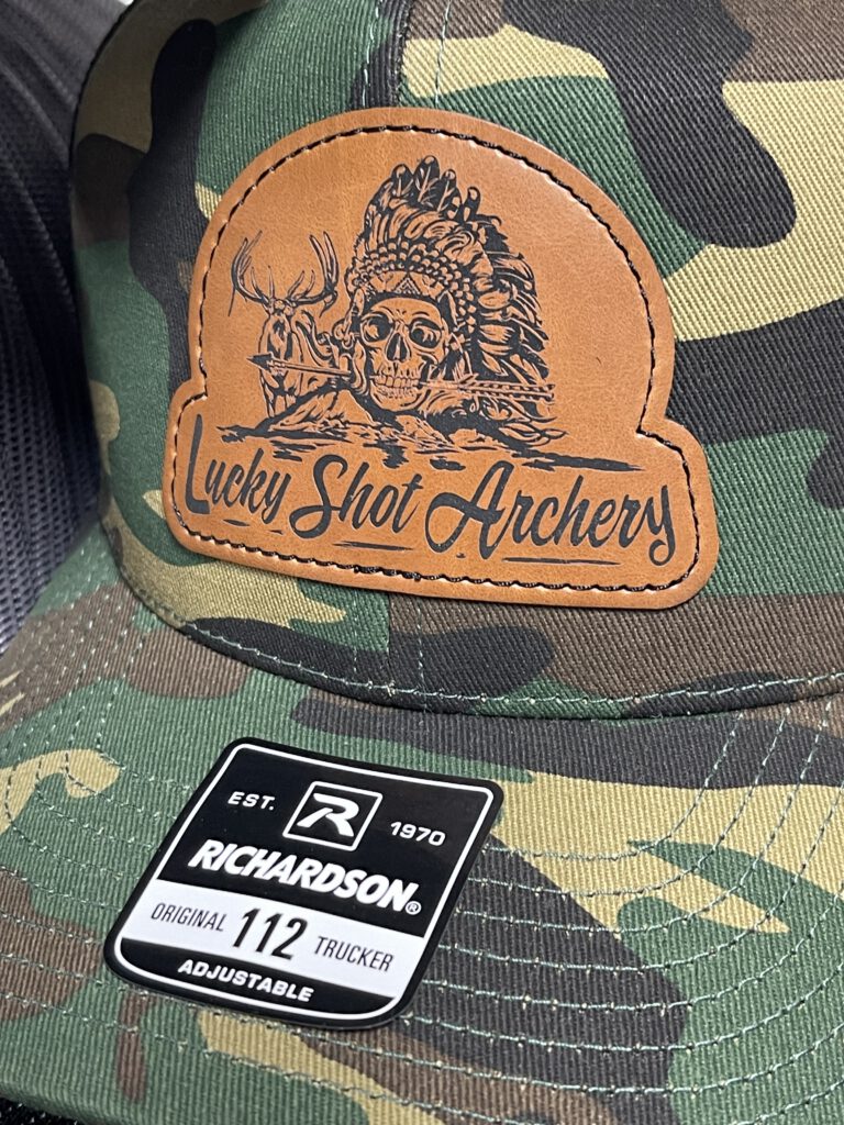 Close-up of a camo trucker hat with a leatherette patch engraved with the Lucky Shot Archery skull, headdress and deer design