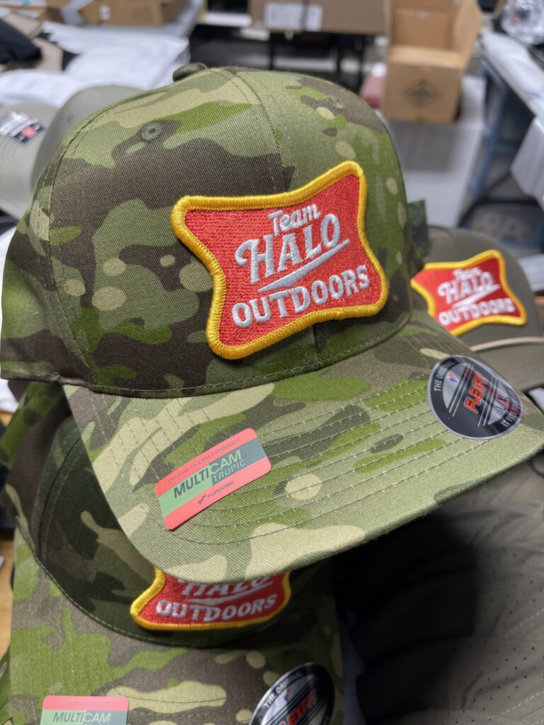 Camo hats with red and gold embroidered Team Halo Outdoors patches, an olive rope hat behind
