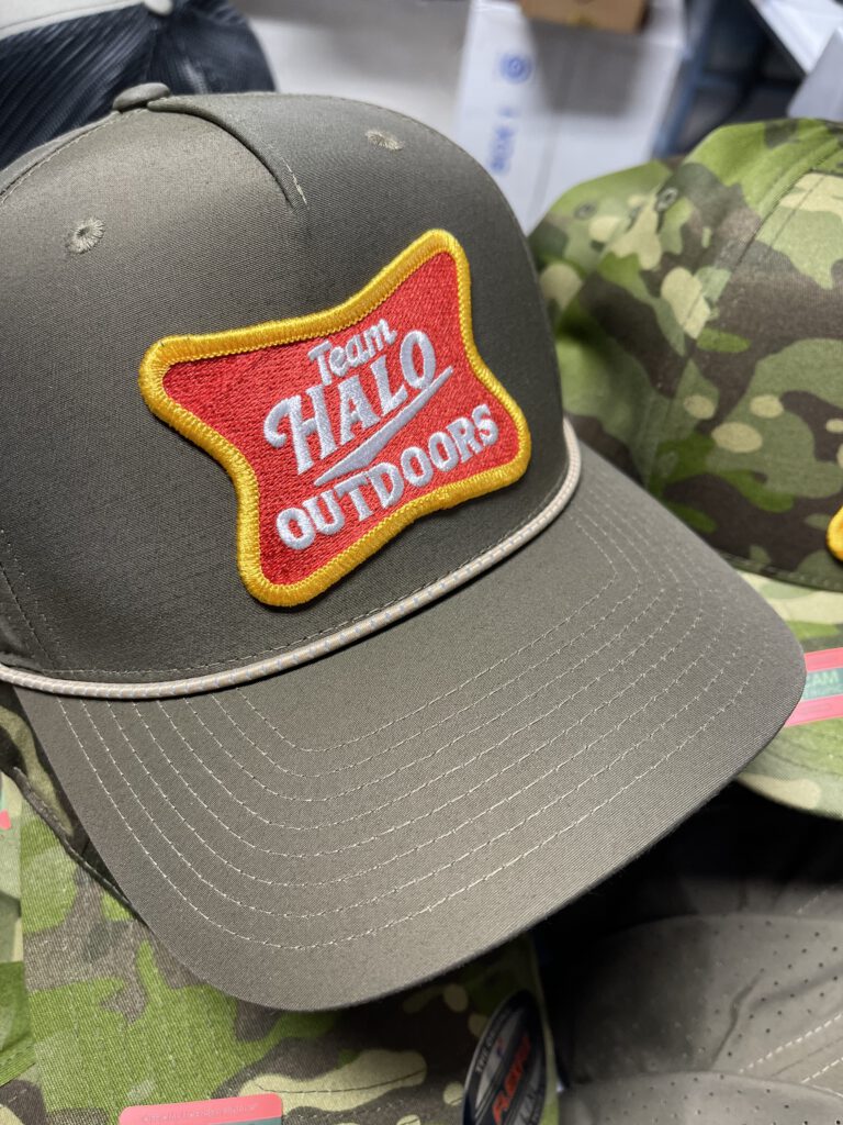 Olive rope hat with a red and gold embroidered Team Halo Outdoors patch, with camo hats behind
