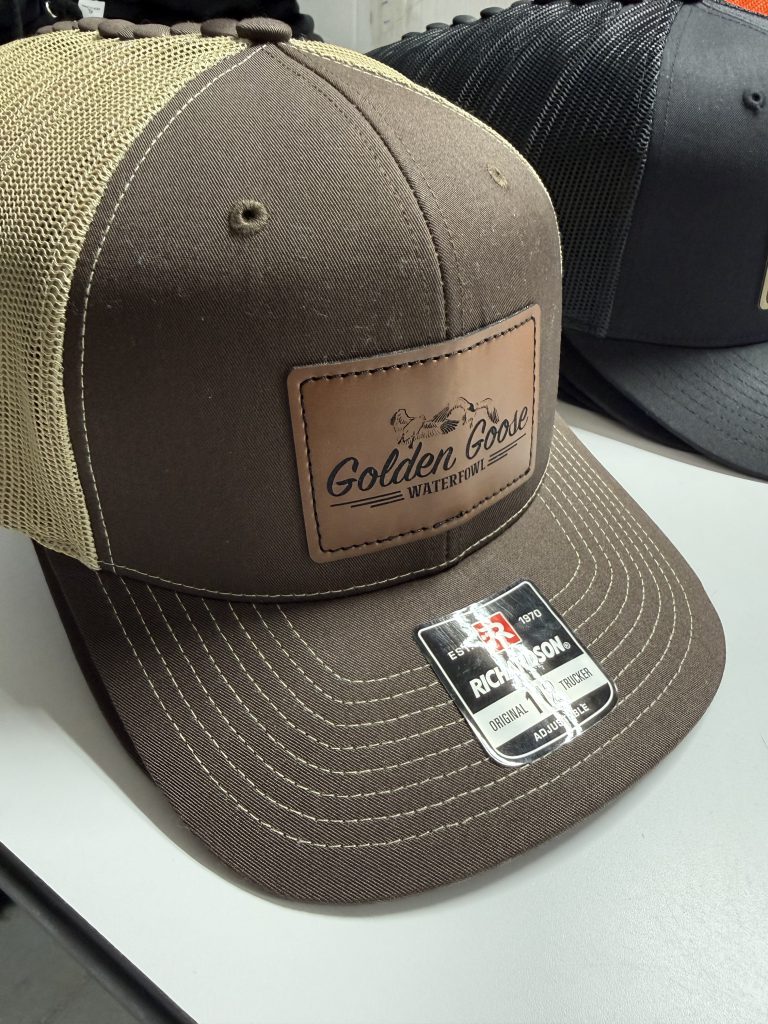 Brown trucker hat with khaki mesh and a brown leatherette patch engraved with the Golden Goose Waterfowl logo