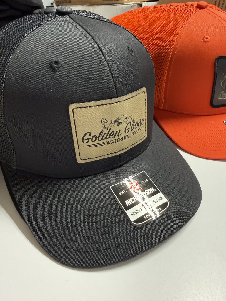 Charcoal trucker hat with a tan leatherette Golden Goose Waterfowl patch, with an orange hat behind it