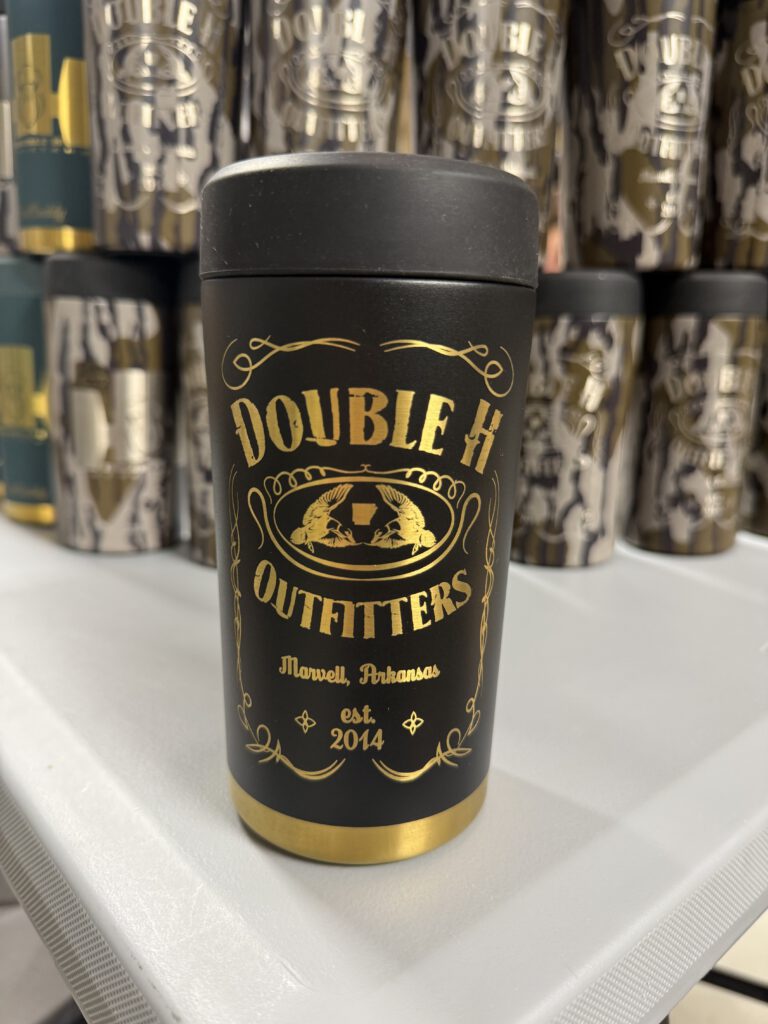 Black can cooler with the Double H Outfitters label design in gold, with camo tumblers behind it