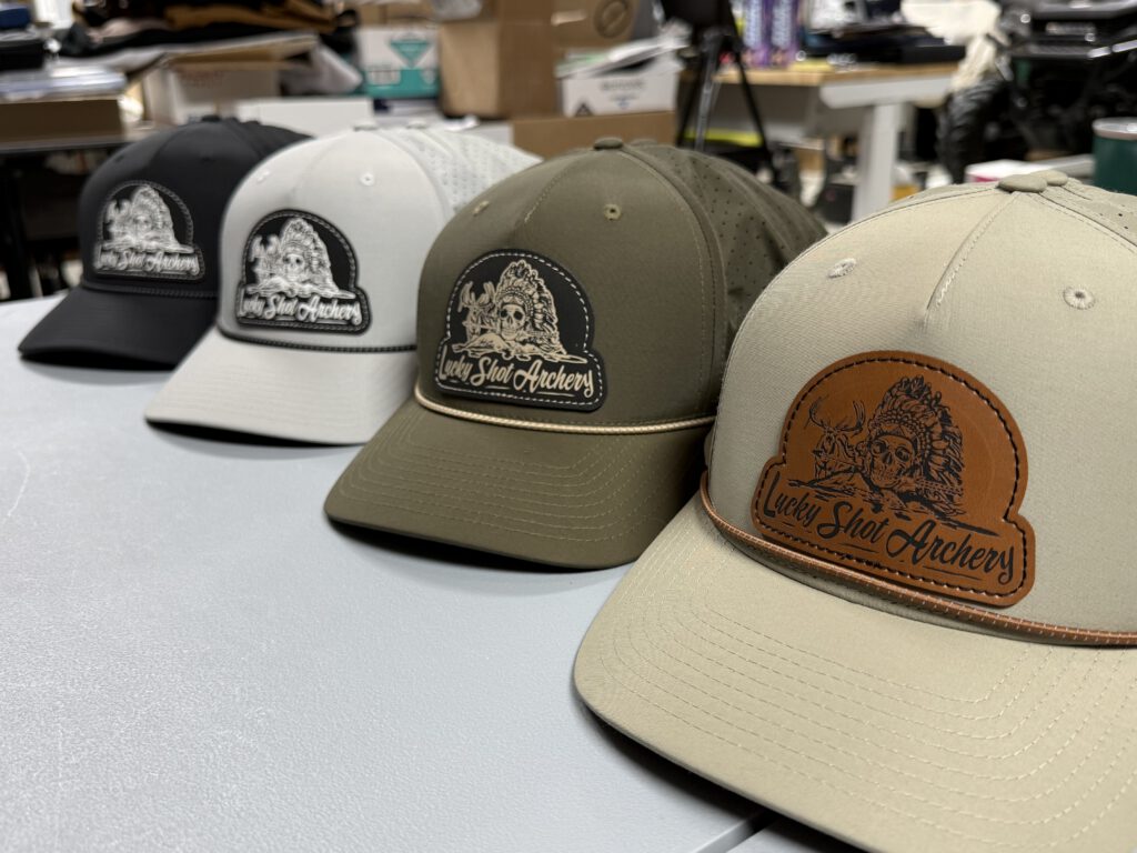 Four rope hats in a row with Lucky Shot Archery patches: black and white patches on black, white and olive hats, and a brown leatherette patch on a tan hat in front