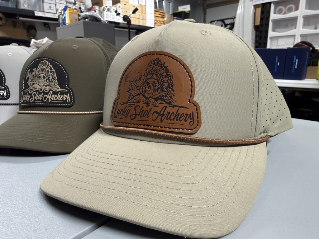 Two rope hats with Lucky Shot Archery leatherette patches, the front one tan with a brown patch engraved with a skull in a feathered headdress