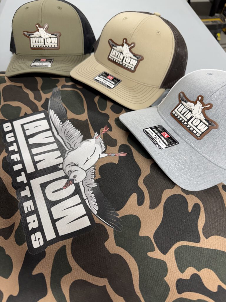 Three trucker hats with Layin Low Outfitters patches resting on a camo shirt printed with the large Layin Low snow goose logo