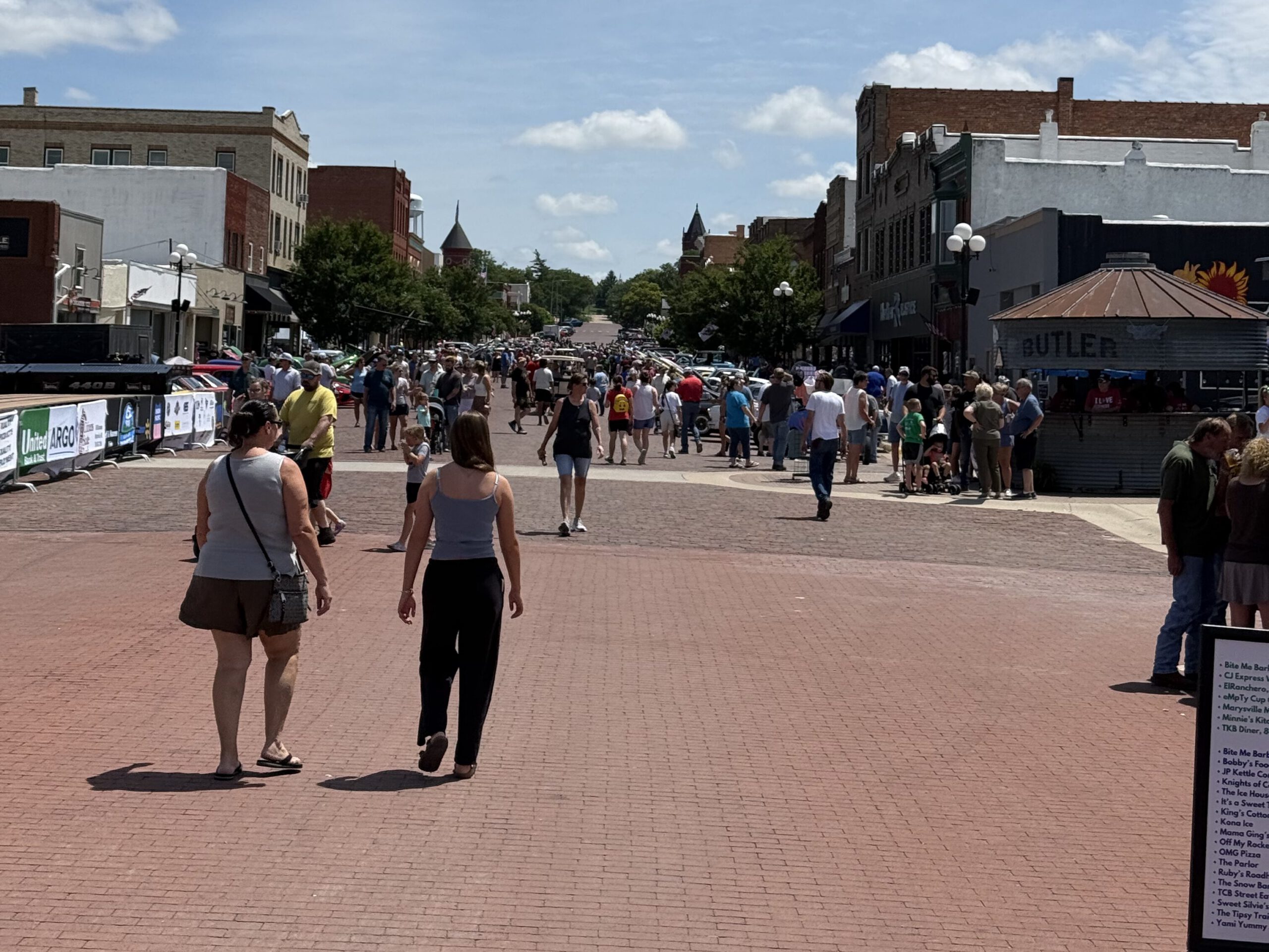 Crowds walking a brick downtown street at a summer event, with storefronts and parked cars beyond