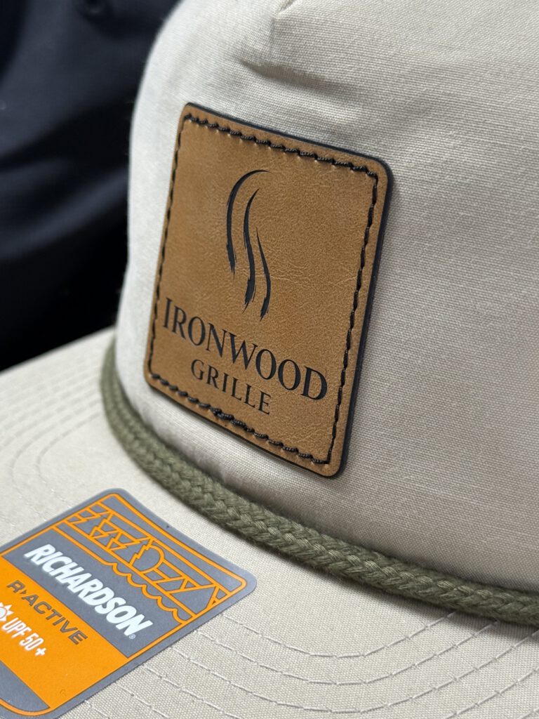 Close-up of a tan leatherette patch with the Ironwood Grille logo on a khaki hat with an olive rope