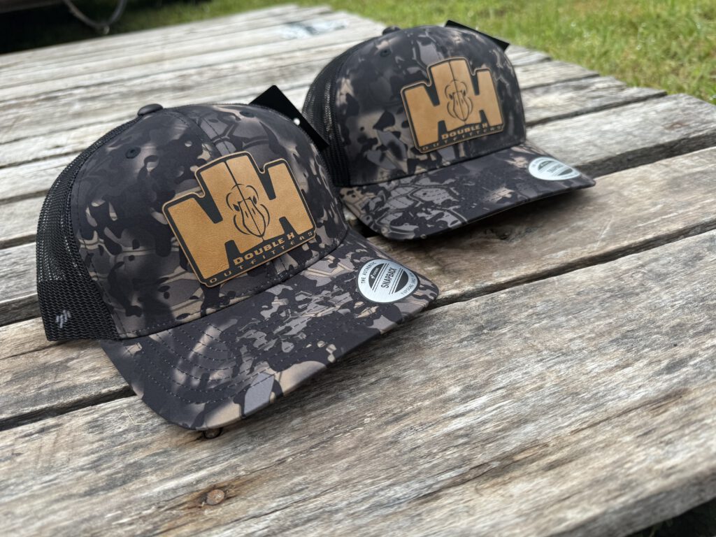 Two dark camo trucker hats with tan leatherette Double H Outfitters patches on a weathered picnic table