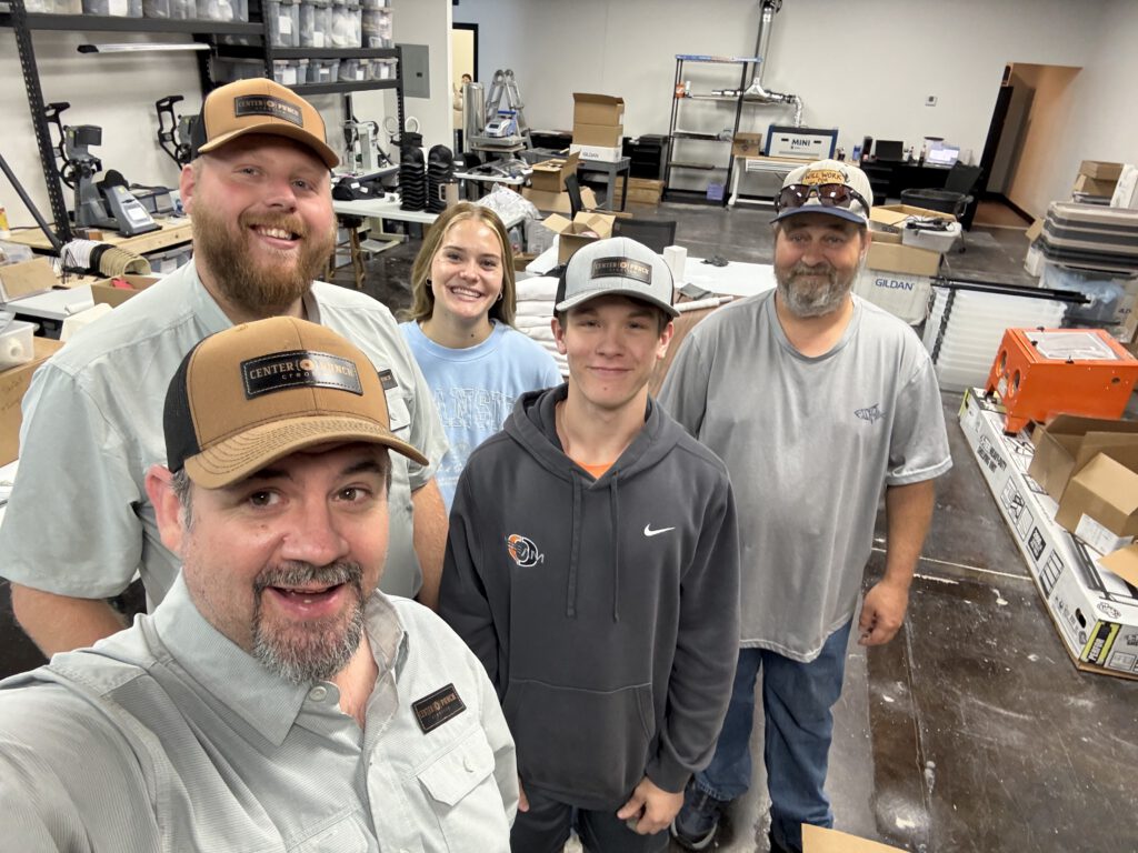 Five members of the Center Punch Creative team smiling together in the shop, most in CPC hats and shirts