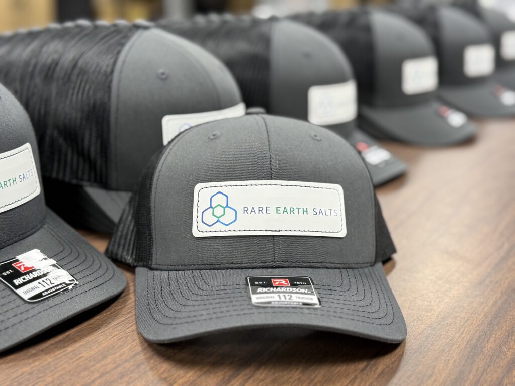 Charcoal and black trucker hats lined up on a table, the front hat showing a white Rare Earth Salts patch with its blue and green hexagon logo