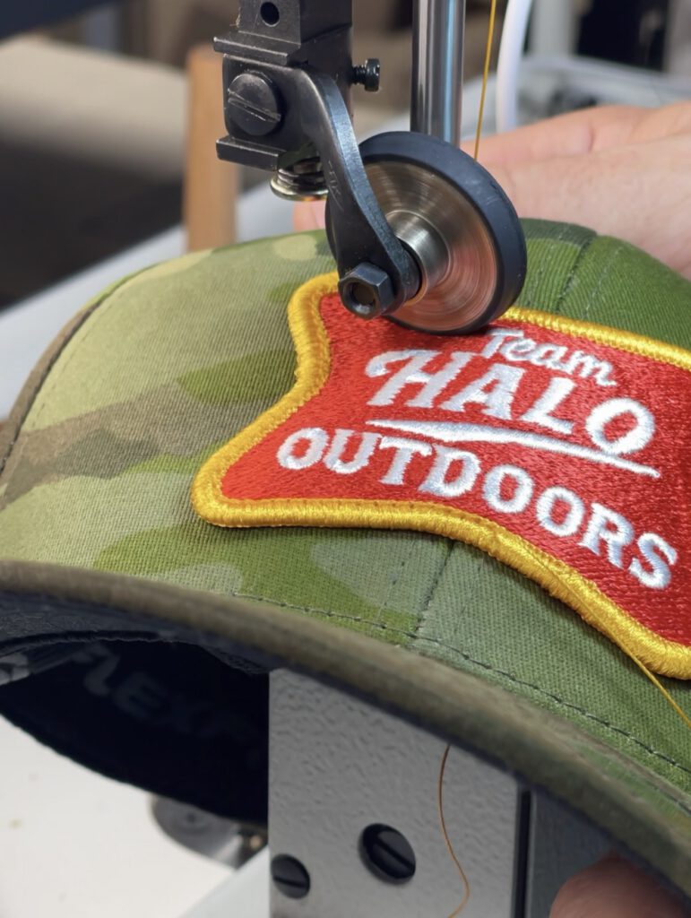 A Team Halo Outdoors patch being sewn onto a camo hat on a post-bed sewing machine, guided by hand