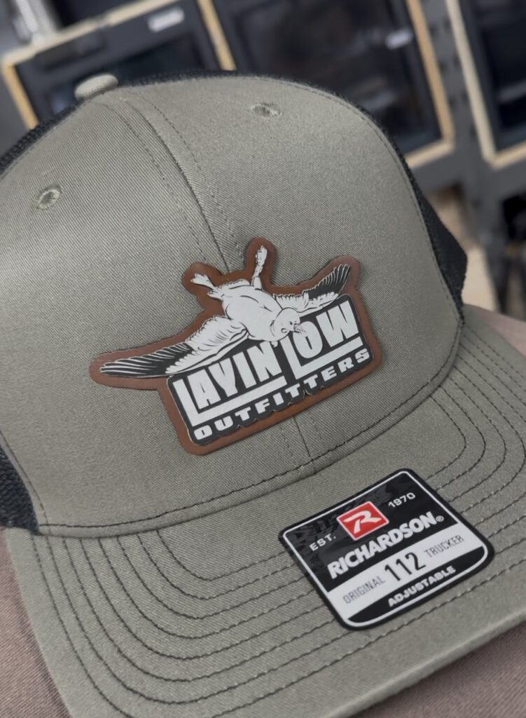 Olive trucker hat with a Layin' Low Outfitters leatherette patch cut to the shape of the logo and its snow goose