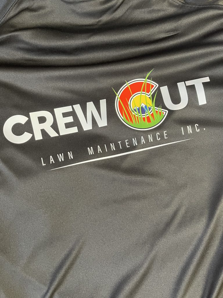 Charcoal performance tee printed with the Crew Cut Lawn Maintenance Inc. logo, the C of Cut drawn as a sun, mountains and grass