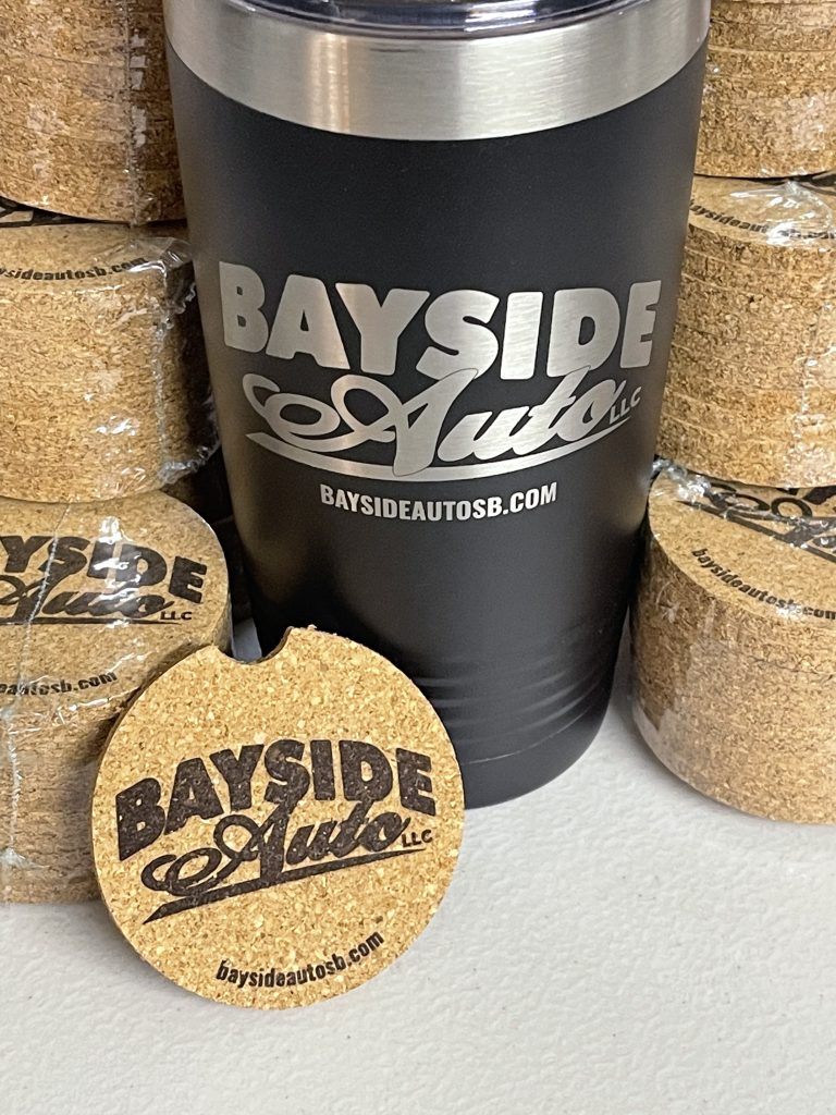 Black tumbler engraved with the Bayside Auto logo, surrounded by stacks of round cork car coasters engraved with the same logo