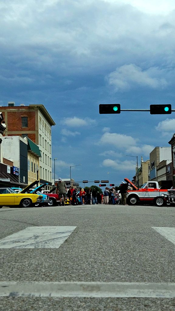 Street-level view of Carfest in downtown Beatrice: classic cars with their hoods up and people walking the show under a cloudy sky