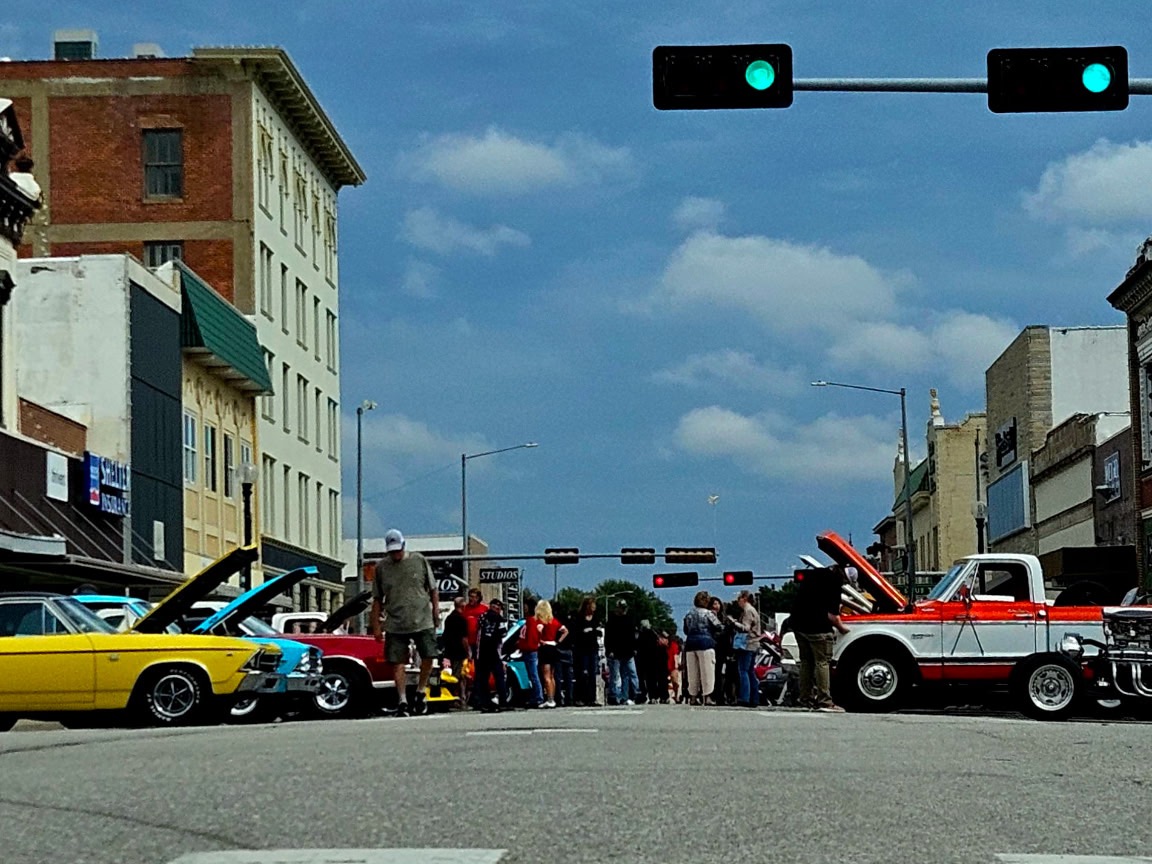 Classic cars with their hoods up and people walking the show on a downtown Beatrice street during Carfest